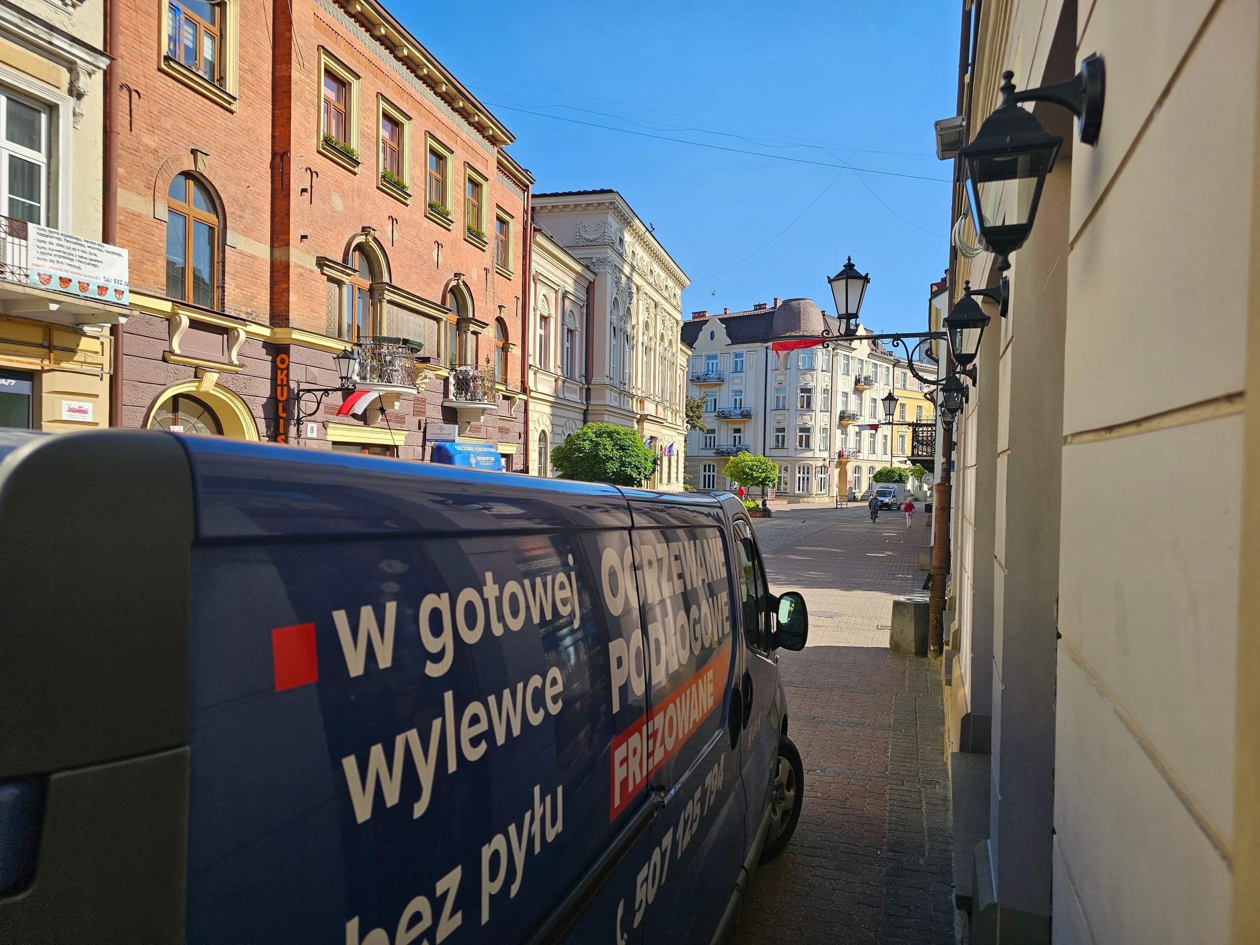 Niebieski bus firmy Kubik z napisem „Ogrzewanie podłogowe frezowane” na rynku miasteczka
